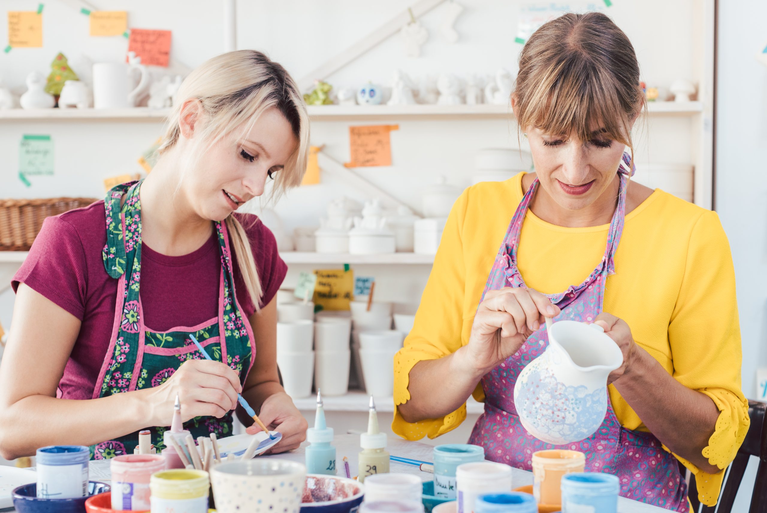 Two women painting own ceramic tableware in DIY workshop with brushes